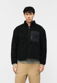 Νεαρός άνδρας που φοράει μαύρο φλις μπουφάν με φερμουάρ και τσέπη στο στήθος με την ετικέτα «GAP» και μπεζ παντελόνι cargo, πάνω σε λευκό φόντο.