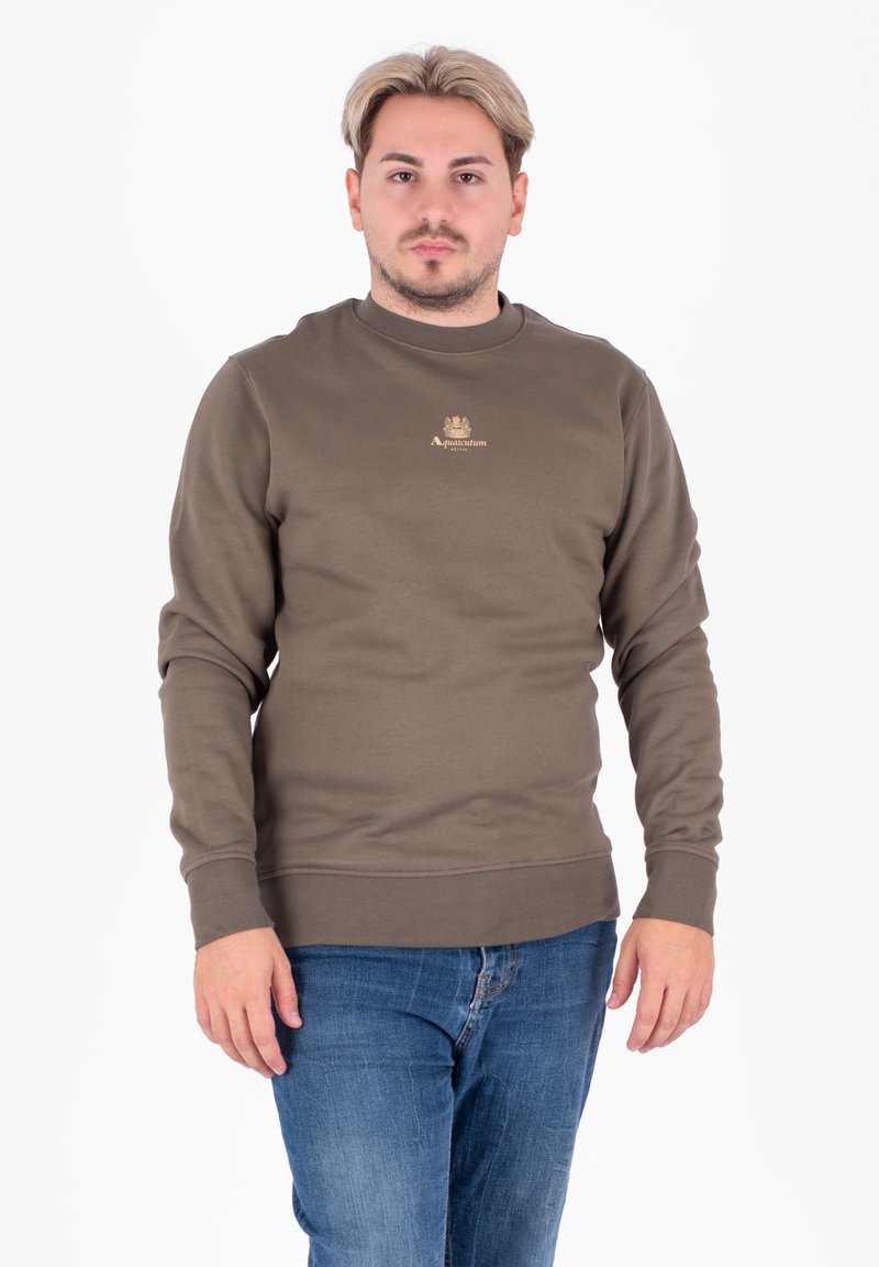 Oliwkowy sweter z miękkiego, teksturowanego materiału. Posiada okrągły dekolt i ściągacze, z małym złotym logo na klatce piersiowej. Noszony z niebieskimi dżinsami.