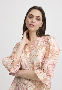Blusa leggera con motivo paisley e maniche a sbuffo, caratterizzata da un scollatura a V. I colori includono rosa tenue, crema e accenti metallici oro.