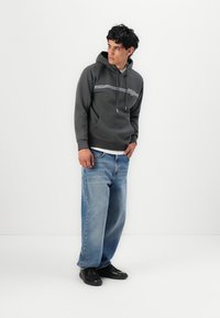 Un sweat à capuche gris avec un cordon de serrage, présenté avec des rayures horizontales bleues et blanches, associé à un jean blue denim ample et des baskets noires.