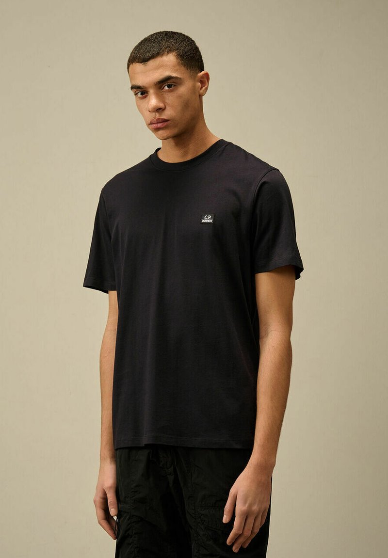 T-shirt in cotone nero con maniche corte, colletto rotondo e piccolo logo sul petto. Tessuto liscio, vestibilità comoda, abbinato a pantaloni corti scuri.