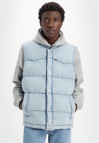 Gilet rembourré en denim bleu clair avec un col, des boutons pression et des poches latérales, porté par-dessus un sweat à capuche gris. Tissu texturé.