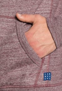 Mano insertada en el bolsillo delantero de un suéter de punto color burdeos con costuras visibles y una pequeña etiqueta de tela azul.