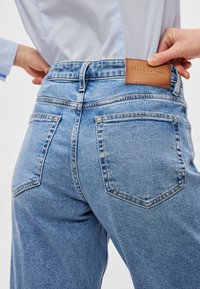 Sinised teksapüksid, millel on kõrge vöökoht, sirge sääreliin ja nähtavad tagataskud. Vöö peal on nahast silt, millel kuvatakse brändi nimi.