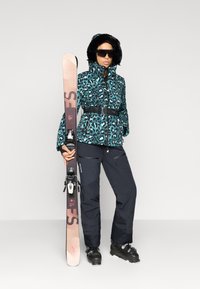 Σαπούνι σκι με τζάκετ σε τυπωμένη ελέφαντα σε τιρκουάζ χρώμα με μαύρη ψεύτικη γούνα στη κουκούλα, συνδυασμένο με μαύρα παντελόνια σκι και μαύρες μπότες σκι, κρατώντας σκι.