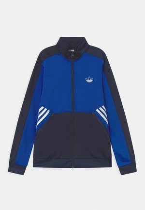 Veste zippée bleue et marine en tissu lisse. Comprend des accents à rayures blanches sur les manches et un petit logo sur la poitrine.