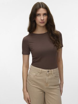 Γυναίκα με μακριά καστανά μαλλιά, που φοράει εφαρμοστή καφέ κοντομάνικη μπλούζα και μπεζ παντελόνι με ψηλή μέση, στέκεται μπροστά σε απλό λευκό φόντο.