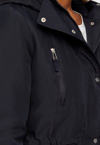 Námořnická modrá bunda s kapucí, přední kapsou na zip, patentním zapínáním a strukturovanou tkaninou, obsahující detail na stažení v pase.