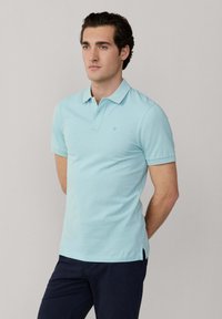 Muž ve světle modré krátkosleevé polokošili a tmavých kalhotách stojí se založenýma rukama za zády na jednobarevném pozadí.