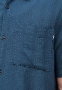 Μπλε κοντομάνικο πουκάμισο με υφή, με τσέπη στο μπροστινό μέρος στο αριστερό στήθος και μικρή υφασμάτινη ετικέτα με το όνομα της μάρκας.