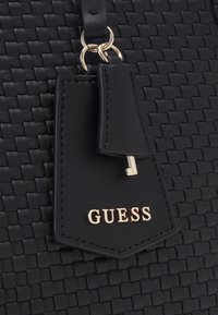 Sac en cuir noir tressé avec étiquette en cuir noir, petite clé en or, anneaux en or, et logo "GUESS" en lettres dorées sur l'étiquette.