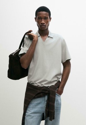 Mladý muž oblečený v světle šedé krátkosvětrové košili, světlých modrých džínách, s hnědým svetrem uvázaným v pase, nesoucí černou tašku přes rameno.