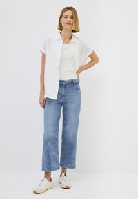 Camisa blanca de lino de manga corta, camiseta de tirantes clara, jeans rectos de color azul claro y zapatillas beige con acentos verdes. Atuendo simple y casual.