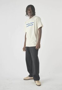 Man with dreadlocks wearing a cream graphic t-shirt, black wide-leg pants, and beige sneakers standing against a plain white background.