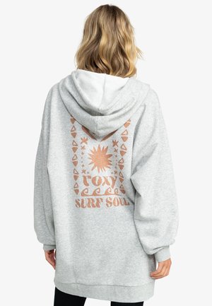 Osoba o blond włosach do ramion, ubrana w jasnoszarą bluzę z kapturem z miedzianym słońcem i napisem „roxy surf soul” na plecach.