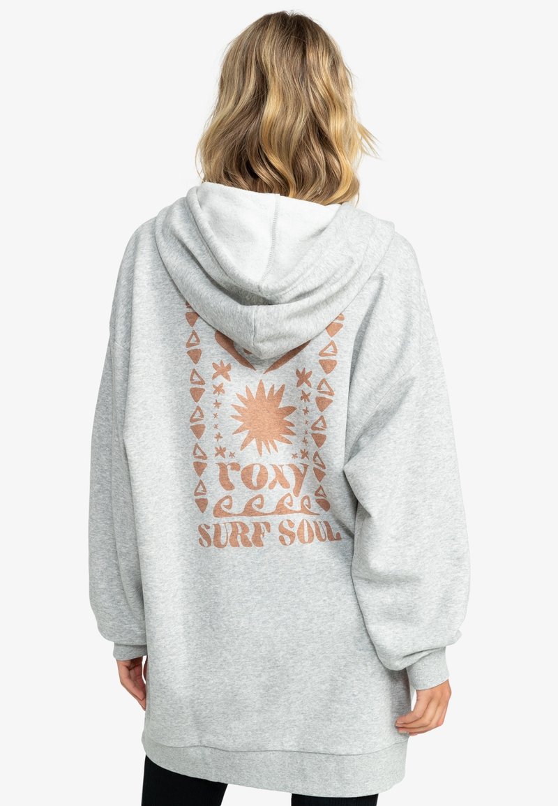 Personne aux cheveux blonds mi-longs portant un sweat à capuche gris clair avec un soleil cuivré et le texte « roxy surf soul » dans le dos.