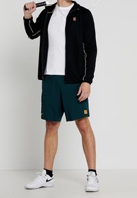 Uomo con giacca nera, t-shirt bianca, pantaloni corti verdi e sneakers bianche che tiene una racchetta da tennis sulle spalle.