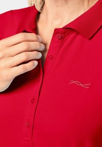 Rotes Polohemd mit Kragen, ausgestattet mit drei Knöpfen und einem kleinen, in hellen Farben gestickten Design. Weicher Baumwollstoff.