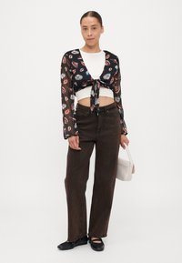 Blusa nera trasparente con stampa di conchiglie colorate, top bianco corto, pantaloni marroni a vita alta e scarpe flat nere. Porta una borsa leggera.