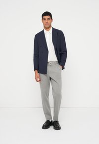 Námořnicky modrý dvouřadý blazer, bílá košile s límečkem, světle šedé kalhoty, černé kožené tenisky, stojící před jednobarevným bílým pozadím.