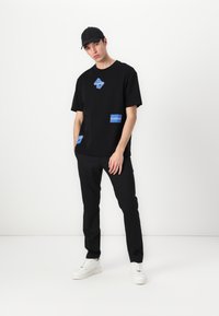 Svart bomulls t-skjorte med blå grafiske lapper, kombinert med svarte slim-fit bukser og hvite joggesko, båret med en svart caps.