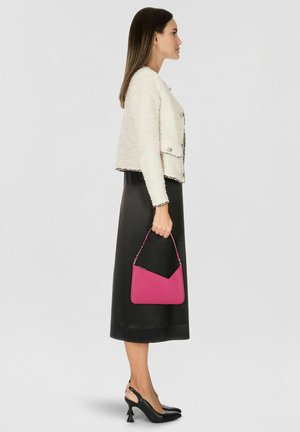 Vrouw in witte, getextureerde jas, zwarte midirok, zwarte hakken, houdt een kleine felroze handtas vast tegen een effen lichte achtergrond, kijkt naar rechts.