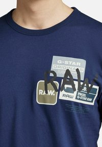 Granatowa koszulka z okrągłym dekoltem; z przodu posiada nadrukowane logo w różnych kolorach (zielony, szary, biały) oraz wyraźny czarny napis "RAW".