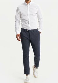 Tmavě modré texturované kalhoty se slim fit, s jedním knoflíkem a bočními kapsami, v kombinaci s bílou košilí na knoflíky a teniskami.