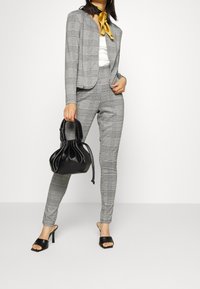Costume en plaid gris avec veste ajustée et pantalon slim, associé à un sac noir et des talons ouverts noirs. Rehaussé d'une écharpe dorée.
