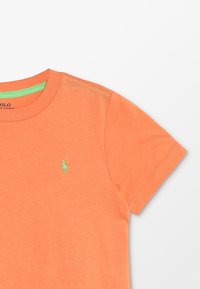 Camiseta de algodón naranja con mangas cortas y cuello redondo. Presenta un acento verde en el cuello y un pequeño logo verde en el pecho.