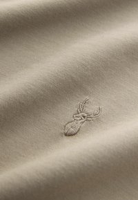 Beige színű pamut anyag finom textúrával, amelyen egy hímzett szarvas embléma látható egy kicsit sötétebb árnyalatban. Sima, puha megjelenés.