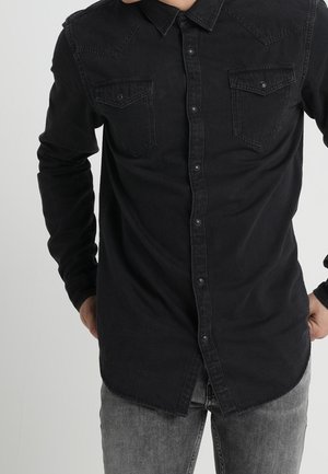 Homme portant une chemise noire boutonnée avec des poches poitrine et un jean gris, debout les mains près des hanches sur un fond uni.
