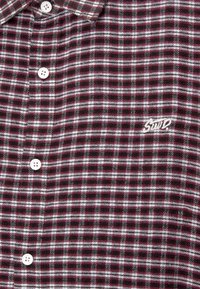 Camicia a quadri in bordeaux, bianco e verde. Realizzata in un tessuto morbido, presenta una chiusura con bottoni e un piccolo logo ricamato.