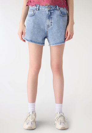 Donna che indossa pantaloncini di jeans azzurri, un top corto di pizzo rosa, calzini bianchi e scarpe da ginnastica bianche, in piedi contro uno sfondo neutro.