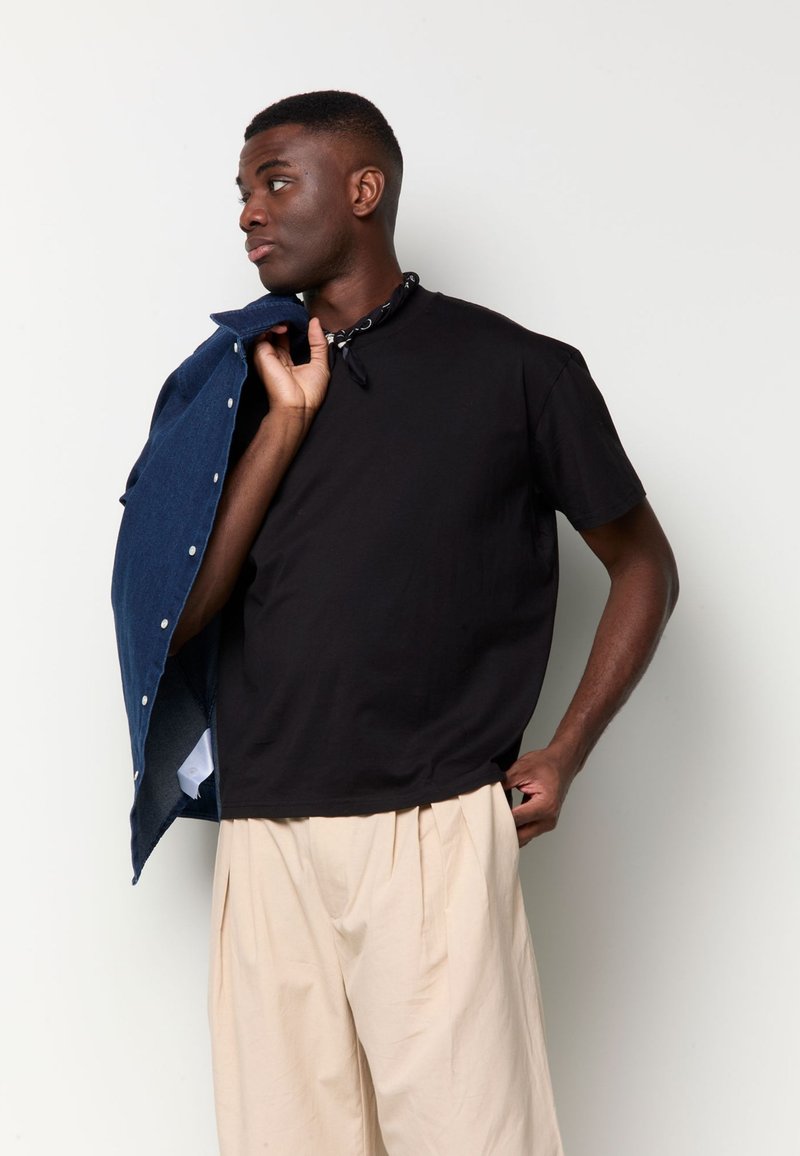 Young man wearing a black t-shirt and beige pants holds a blue jacket over his shoulder, looking to his left against a plain background.