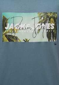 Tissu bleu avec un motif rectangulaire de palmiers sur fond de ciel et un texte partiellement visible indiquant « Jack's Jeans Denim Since Ninete Ninety ».