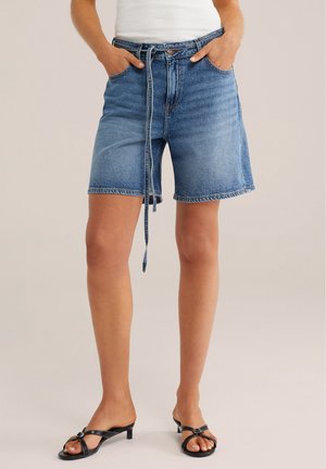 Persona con pantalones cortos de mezclilla azul hasta la mitad del muslo con cinturón anudado al frente, camiseta blanca sin mangas y sandalias negras de tacón abiertas en los dedos, de pie con las manos en los bolsillos.