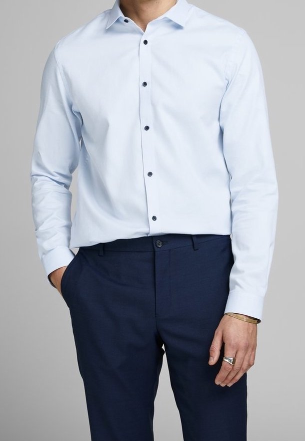 Man som bär en ljusblå långärmad skjorta med knappar och mörkblå byxor, vänster hand i fickan, höger hand visar en silverring och armband.