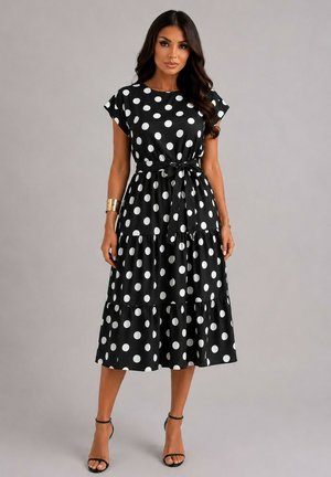 Femme portant une robe midi noire à pois blancs, manches courtes, ceinture à la taille et talons noirs à bride à la cheville, debout devant un fond uni.