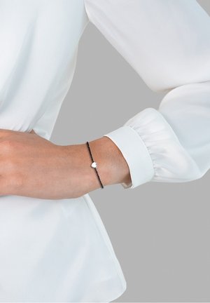 Personne portant un délicat bracelet chaîne noir avec un petit pendentif en forme de cœur en argent au poignet, vêtue d'une chemise blanche à manches longues.