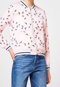 Růžová bomber bunda s vzorem černých vlaštovek, se zapínáním na zip a pruhovanými manžetami a lemem v modré a bílé barvě. Hladká textura látky.