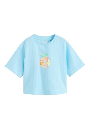 Ung pige smiler iført en lyseblå oversized skjorte med orange design, denimnederdel og farverige hæklede sandaler.