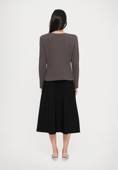 Woman facing away wearing a textured brown blazer, black midi skirt, and white kitten heels against a white background.