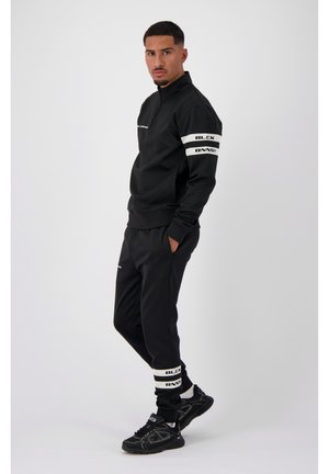 Zwart sportief trainingspak met witte logo-accenten. Het jack met rits heeft lange mouwen, terwijl de broek elastische boorden heeft met merkopdruk. Zwarte sneakers.