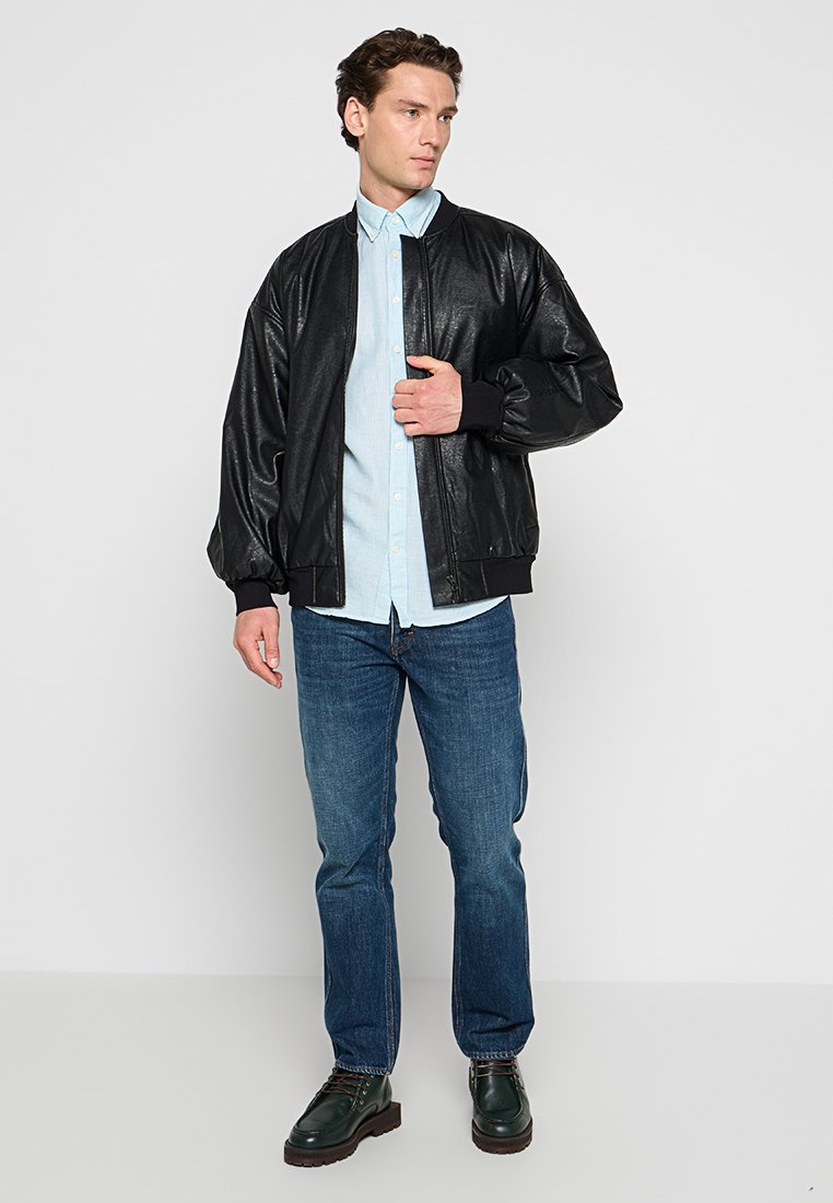 Ein junger Mann trägt eine schwarze Bomberjacke über einem hellblauen Hemd, blaue Jeans und dunkelgrüne Stiefel, während er vor einem einfarbigen Hintergrund steht.