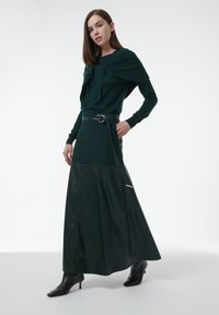 Lange groene gebreide top met gedrapeerde details, gecombineerd met een donkergroene leren maxi-rok met een zijrits en een zwarte riem. Zwarte haklaarzen.