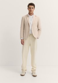 Beige strukturiertes Blazer mit zwei Taschen, weißem Hemd darunter, hellbeigen Hosen und cremefarbenen Sneakers. Neutrale Farbpalette insgesamt.
