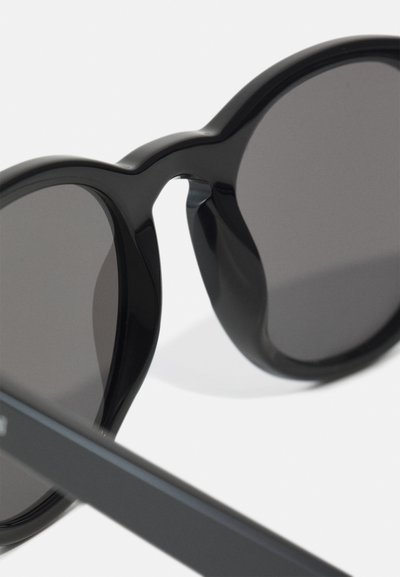 Occhiali da sole neri con lenti oversize e tonde, finitura lucida. Presentano una montatura ampia e tempie nere sottili, offrendo un design elegante.