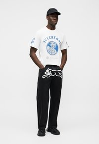 Λευκό κοντομάνικο μπλουζάκι με μπλε γραφικό παγωτού και κείμενο, συνδυασμένο με μαύρο παντελόνι που διαθέτει σχέδιο με λευκή περίγραμμα γάτας, φορεμένο με μαύρα αθλητικά παπούτσια.