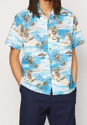 Homme portant une chemise hawaïenne bleue à manches courtes avec un motif de palmiers, bateaux et océan, associée à un pantalon bleu foncé, mains dans les poches.